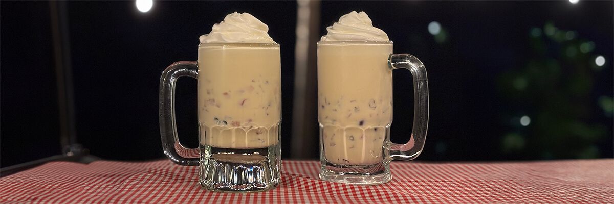 Two large glass mugs filled with a creamy, pale-colored dessert drink containing small colorful mix-ins and topped with a swirl of whipped cream sit side by side on a red-and-white checkered tablecloth at night. Blurred lights glow in the background.