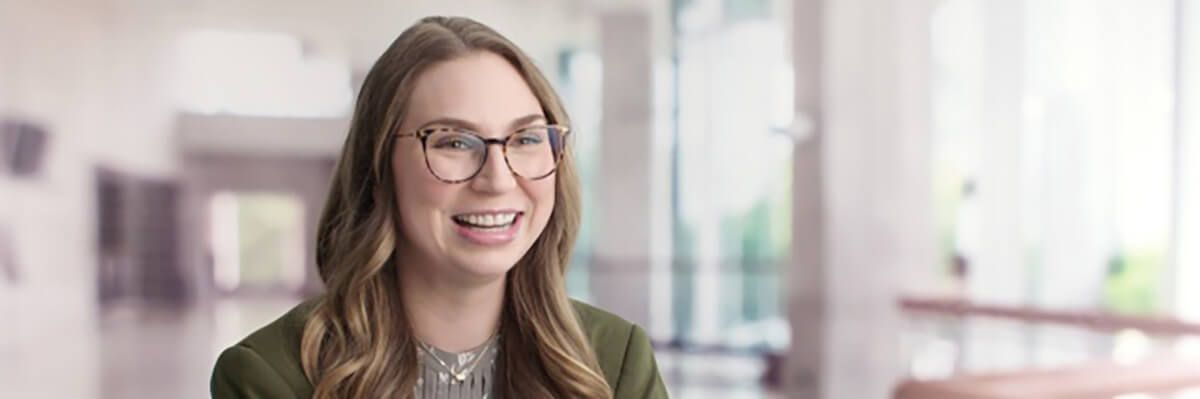 A woman with long, light-brown hair, glasses, and a grey sweater under an olive-green blazer is pictured from the shoulders up. She sits in the left side of the frame and is smiling, looking just to the right of the camera photographing her. She is pictured in a bright hallway with white walls and floors and a wall of windows.
