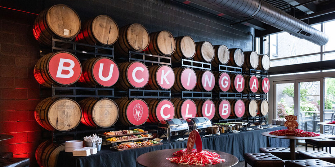 A buffet setup in front of a wall of wooden barrels, each marked with red circular signs spelling "BUCKINGHAM CLUB"; the room features industrial-style decor with exposed ductwork, large windows, and round tables with red decorations.
