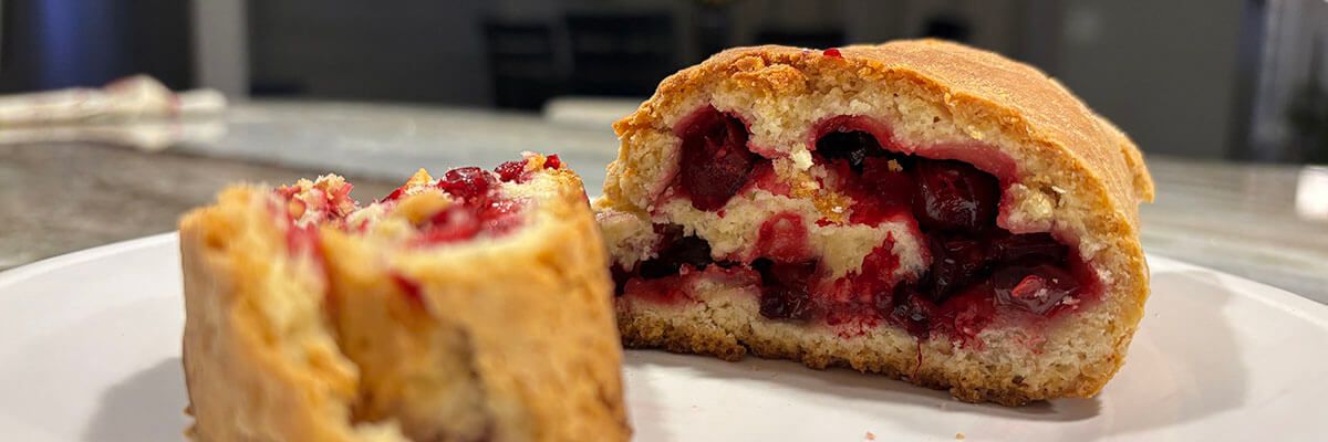 A golden brown, log-shaped baked good sits on a white plate. The baked good is cut, revealing a cross-section that features red berries in the center.