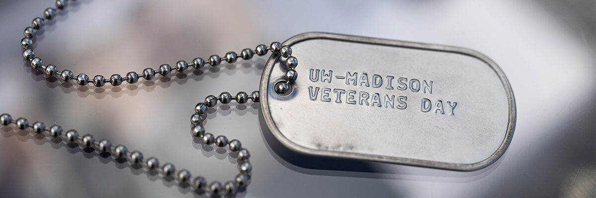 A close-up shot of a silver dog tag on a silver chain. The dog tag reads "UW-Madison Veterans Day."