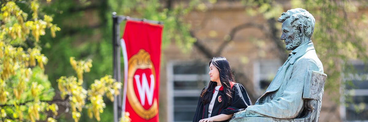 A woman with long dark hair is sitting on the lap of a green statue of a seated man. Both are pictured in profile, looking off to the left of the frame. The woman is wearing a black gown with red accents over a white top. In the left of the background, a red, vertical banner features a crest logo with a white, serif W in the middle. The rest of the background appears to be an academic building of light brick.