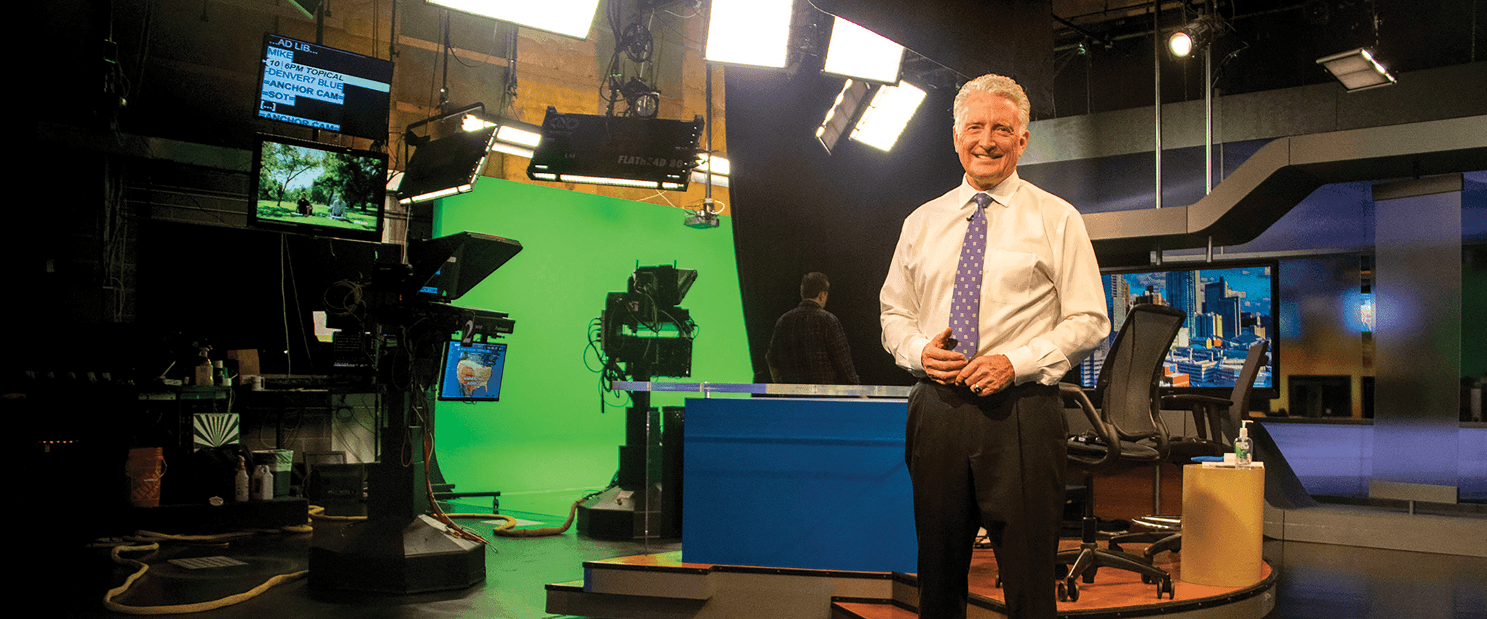 A television news studio with bright overhead lights, cameras, and a green screen backdrop. In the foreground, a smiling male news anchor in a white shirt, purple tie, and black slacks stands near a desk.
