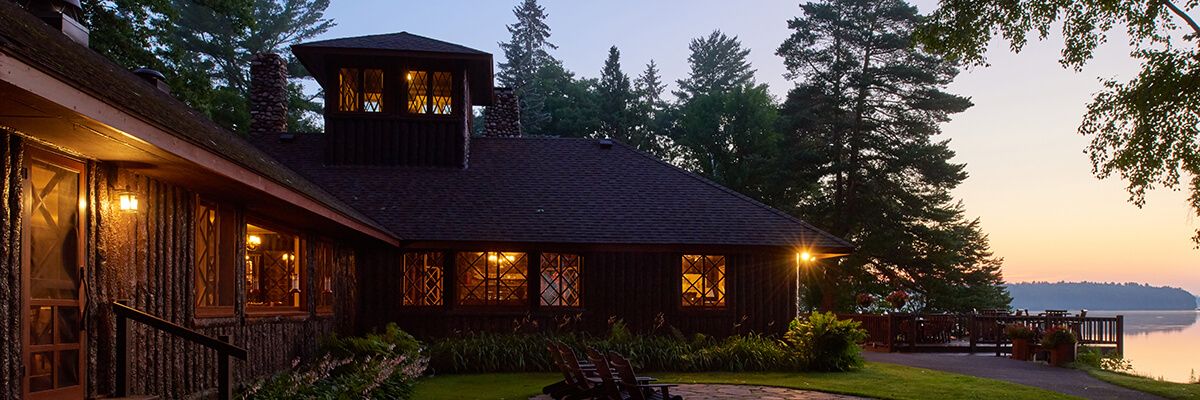 Rustic lakeside lodge at dusk, with warm lights glowing through diamond-pane windows. The dark wood building is surrounded by tall pine trees and landscaped greenery, with Adirondack chairs on a stone patio and a deck overlooking calm water in the background.