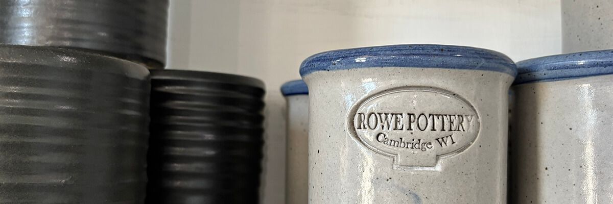 Stoneware crocks with blue rims on a shelf, featuring an embossed "Rowe Pottery, Cambridge WI" logo.